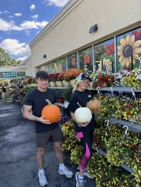 Two staff members holding pumpkins outside the Garden Center entrance, beside racks of hanging flower baskets