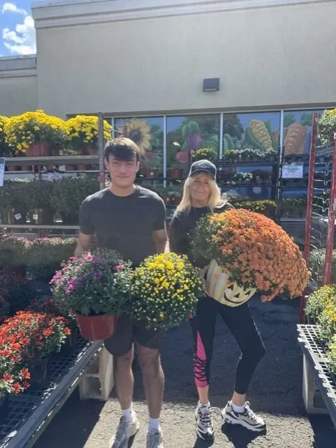 Two staff members carrying mums and a pumpkin planter outside the garden centre