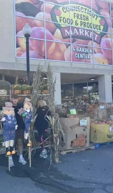 Two staff members among scarecrows and corn stalks beneath the Connecticut Fresh Food & Produce Market storefront sign